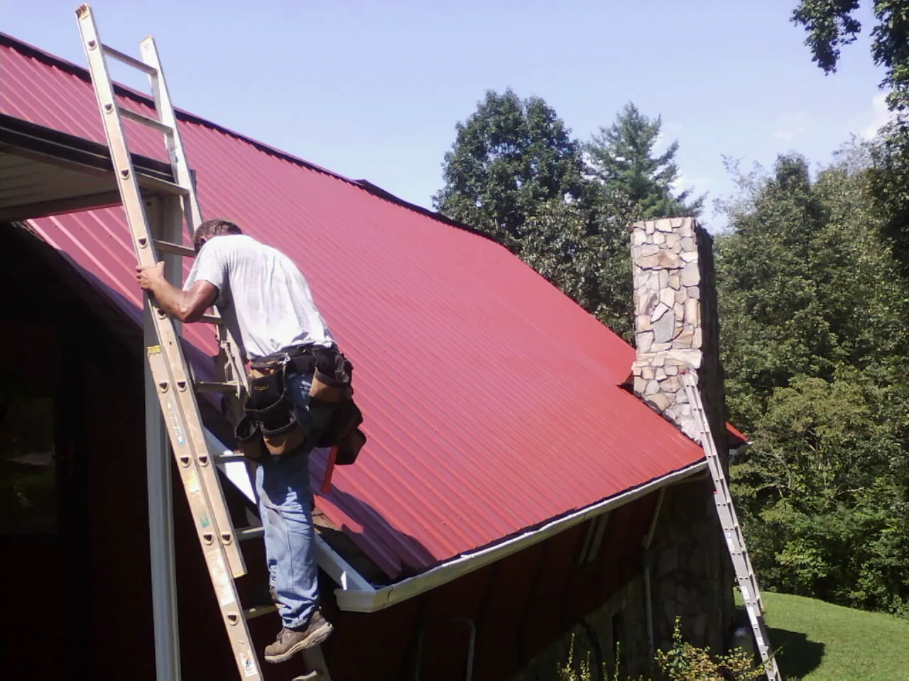 Roofer on a ladder beside a red metal roof and stone chimney — Soffit & Fascia Repair by Roofing Brothers in Tarrytown